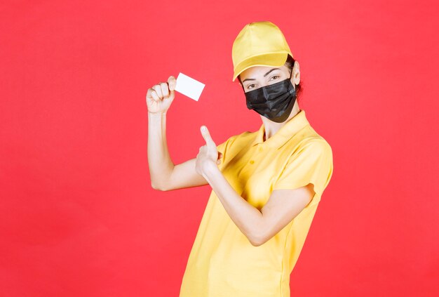 Female courier in yellow uniform and black mask presenting her business card and showing thumb up