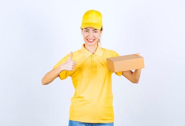 Женский курьер в желтой форме доставляет картонную посылку и показывает знак успешной руки.