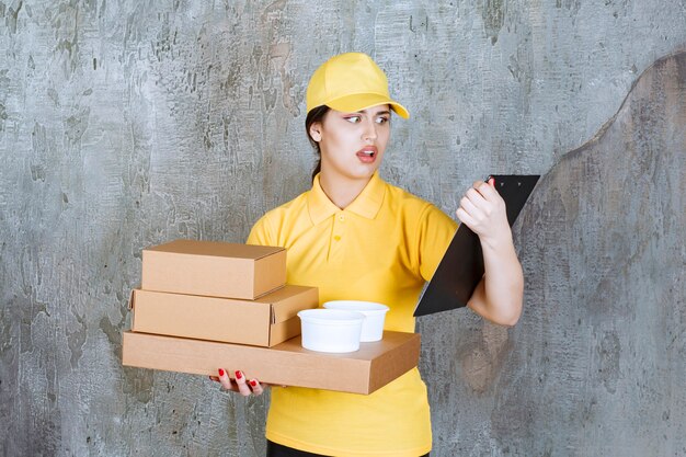 Female courier in yellow uniform delivering multiple cardboard boxes and takeaway cups and checking the address on the list.