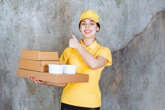 Female courier in yellow uniform delivering multiple cardboard boxes and takeaway cups and showing positive hand sign