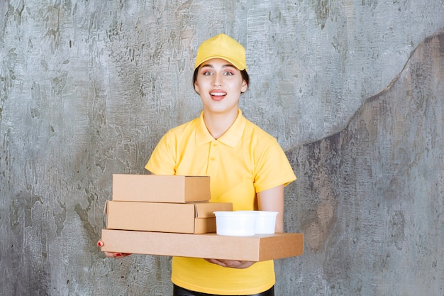 Female courier in yellow uniform delivering multiple cardboard boxes and takeaway cups.