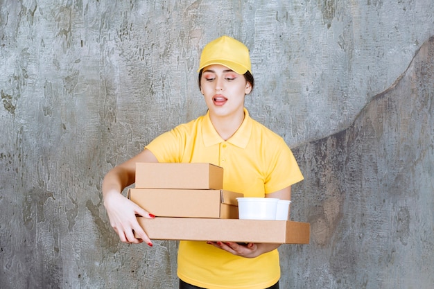 Female courier in yellow uniform delivering multiple cardboard boxes and takeaway cups