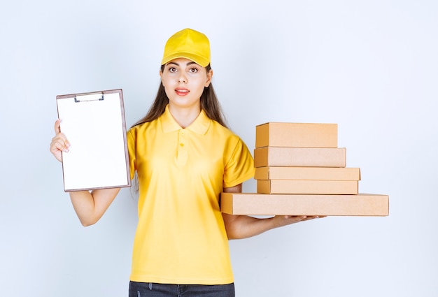 Female courier in yellow uniform holding bunch of cardboard boxes and clipboard over white wall.