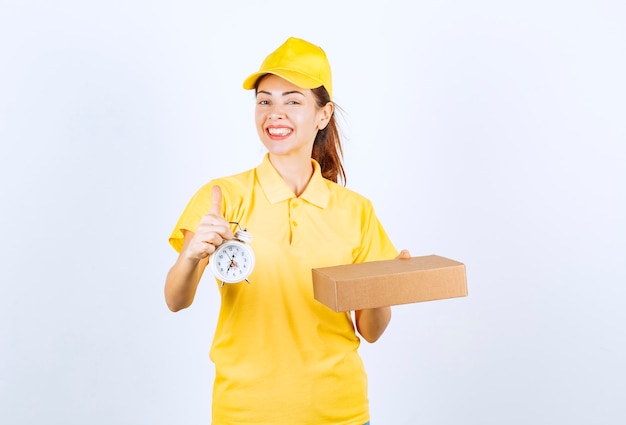 Corriere femminile in uniforme gialla che tiene una scatola di cartone e una sveglia che significa la consegna espressa in tempo.