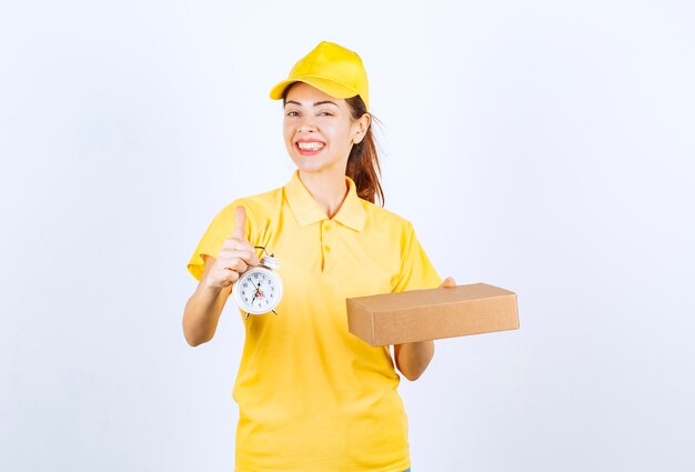 노란색 제복을 입은 여성 택배기사가 판지 상자와 제시간에 신속하게 배달된다는 알람 시계를 들고 있습니다.