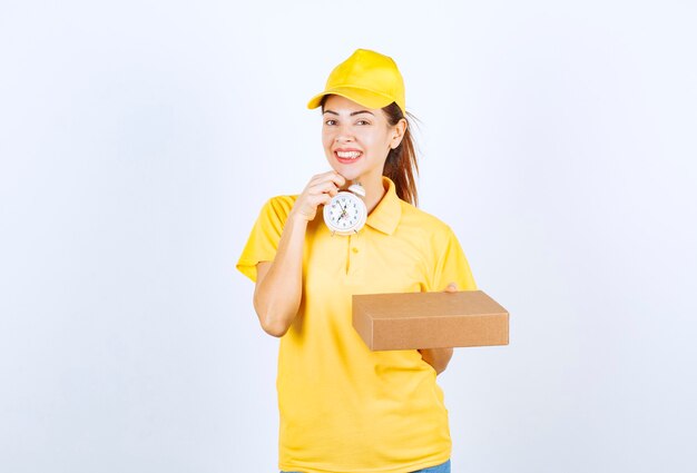 Женский курьер в желтой форме держит картонную коробку и будильник, что означает своевременную экспресс-доставку.