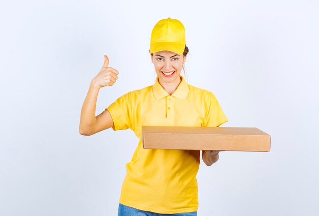 Female courier in yellow uniform holding a cardboard parcel and showing enjoyment hand sign. 
