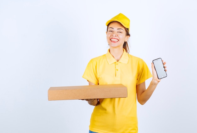 Female courier in yellow uniform holding a cardboard parcel and showing her white smartphone. 