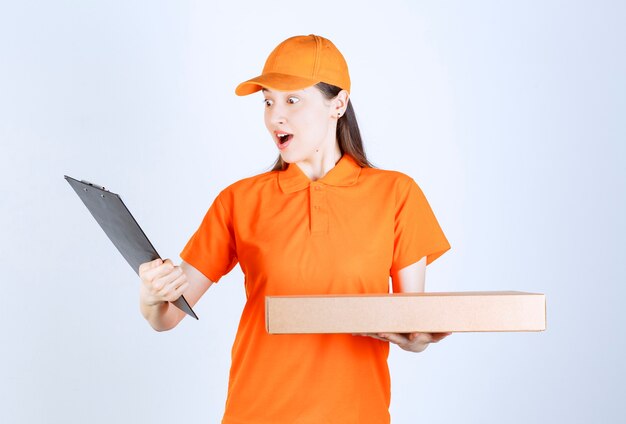 Female courier in yellow uniform holding a cardboard takeaway box and checking the address list and looks confused.