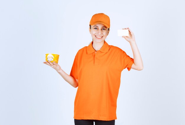 Female courier in yellow uniform holding a takeaway noodles cup and presenting her business card.