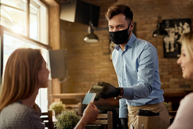 Female customer making contactless payment to a waiter who is wearing protective face mask in a cafe