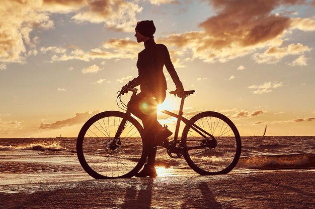 그녀의 자전거와 함께 서 있고 바다 해안에서 일몰을 즐기는 여성 사이클.