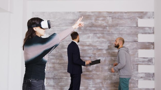 Female designer with virtual reality goggles in empty apartment while real estate agent is talking with the homeowner in the background.