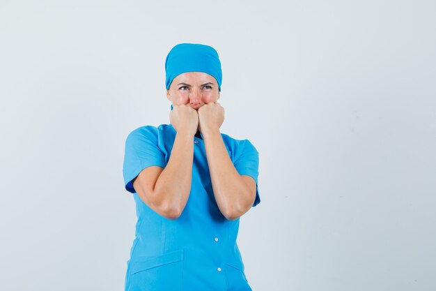 Female doctor biting fists emotionally in blue uniform and looking scared , front view.