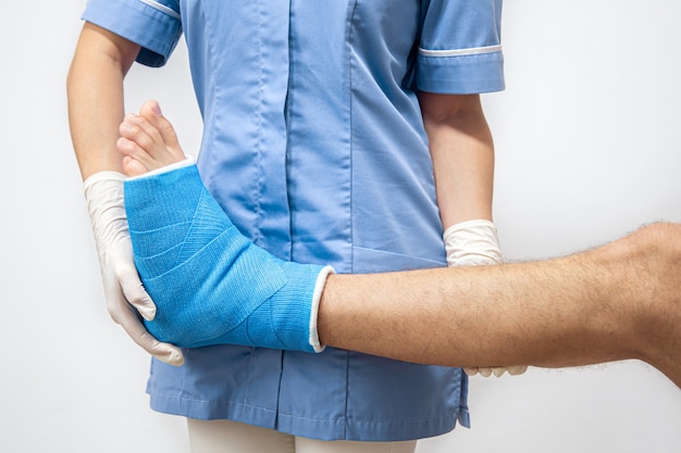 Female doctor in a blue medical gown checking broken leg on male patient.