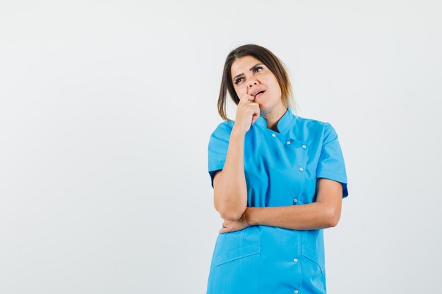 Женщина-врач в синей форме кусает палец и смотрит задумчиво