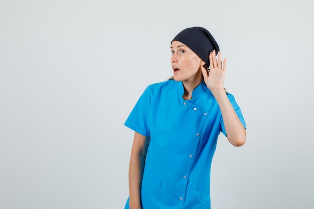 Female doctor in blue uniform, black hat holding hand behind ear to listen and looking focused