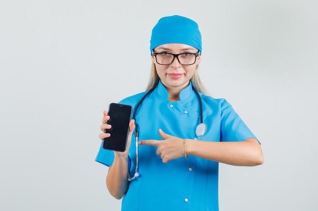 Female doctor in blue uniform, glasses pointing finger at smartphone and looking cheerful