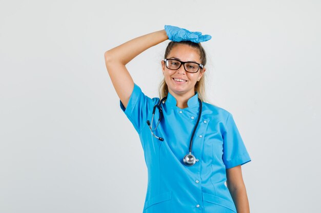 Женщина-врач в синей форме, перчатках, очках поднимает руку с рукой на голове и выглядит весело