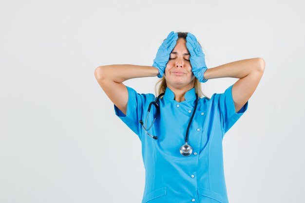 Female doctor in blue uniform, gloves holding head with hands and looking regretful