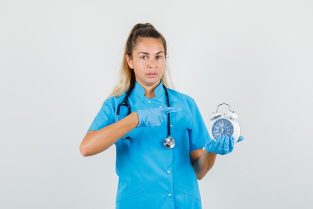 Female doctor in blue uniform, gloves pointing finger at alarm clock