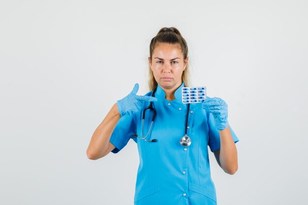 Female doctor in blue uniform, gloves pointing at pack of capsules and looking serious