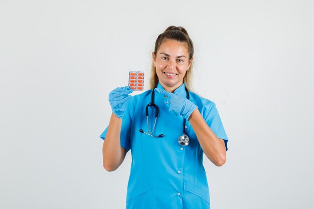 Female doctor in blue uniform, gloves pointing at pack of pills and looking cheery