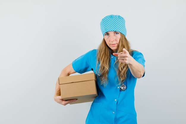 Female doctor in blue uniform holding cardboard box and pointing at camera 