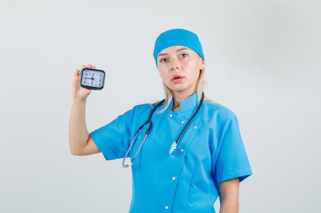 Женщина-врач в синей форме держит часы и выглядит пунктуально