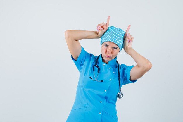 Женщина-врач в синей форме держит пальцы над головой как бычьи рога и выглядит смешно, вид спереди.