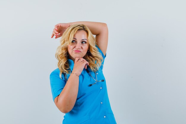 Female doctor in blue uniform holding hand on head while posing and looking self-confident