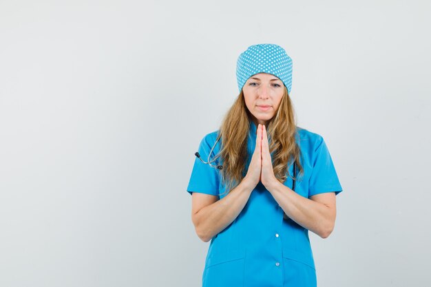 Женщина-врач в синей форме, взявшись за руки в молитвенном жесте и выглядя с надеждой