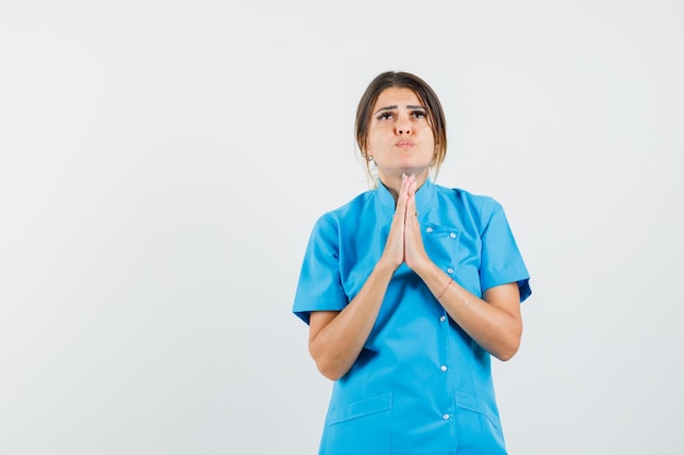 Женщина-врач в синей форме, взявшись за руки в молитвенном жесте и выглядя с надеждой