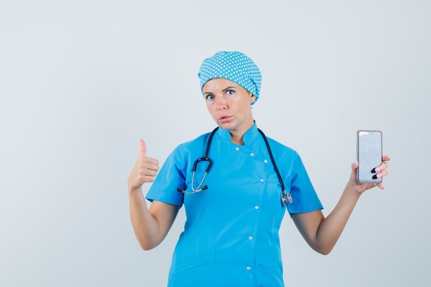 파란색 제복을 입은 여성 의사가 휴대 전화를 들고 엄지 손가락을 보여주는 전면 뷰.