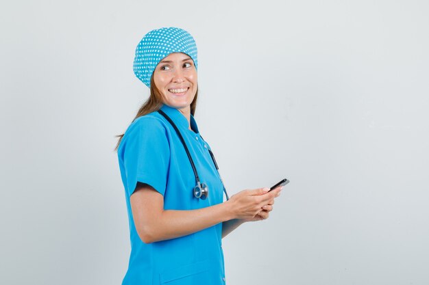 Женщина-врач в синей форме, глядя, держа смартфон и выглядя счастливой.