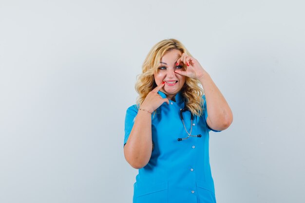 Female doctor in blue uniform making round on eye while pointing at her teeth and looking funny space for text