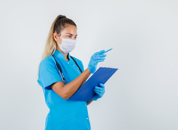 Female doctor in blue uniform, mask, gloves pointing pencil away while holding clipboard