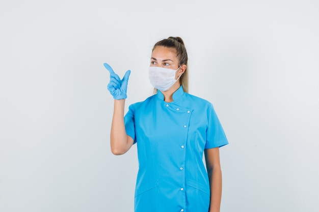 파란색 유니폼, 마스크, 장갑 총 제스처로 측면을 가리키고 초점을 맞춘 여성 의사