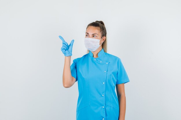 Женщина-врач в синей форме, маске, перчатках указывает в сторону жестом пистолета и выглядит сосредоточенным