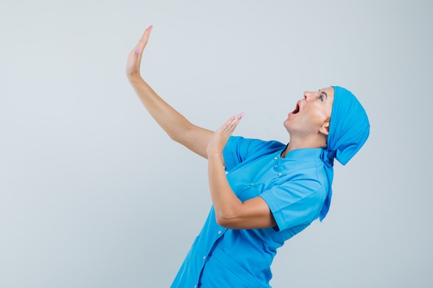 Dottoressa in uniforme blu alzando le mani per difendersi e guardando spaventato, vista frontale.