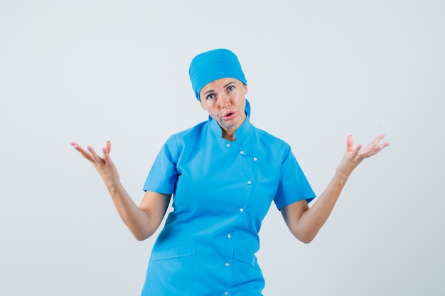 Женщина-врач в синей форме вопросительно поднимает руки и выглядит озадаченно, вид спереди.