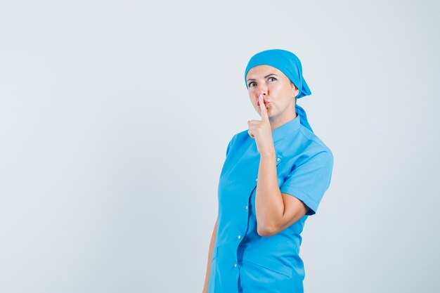沈黙のジェスチャーを示し、注意深く見ている青い制服を着た女性医師、正面図。