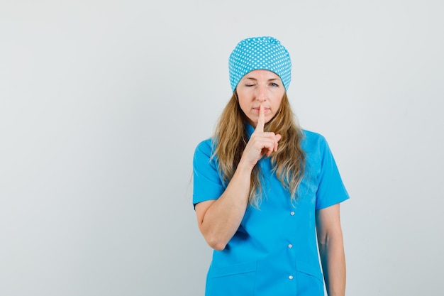 Female doctor in blue uniform showing silence gesture and winking eye 
