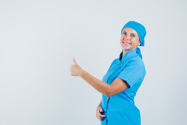 Женщина-врач в синей форме показывает палец вверх и выглядит радостным, вид спереди.