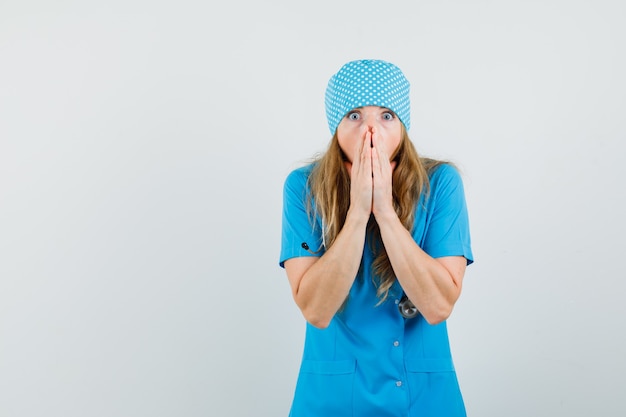 Женщина-врач, сжимая руки возле открытого рта в синей форме и выглядя шокированной.