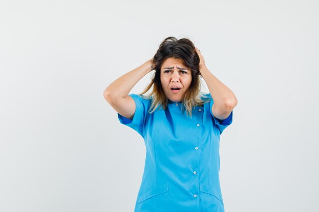 Female doctor clasping head with hands in blue uniform and looking distressed