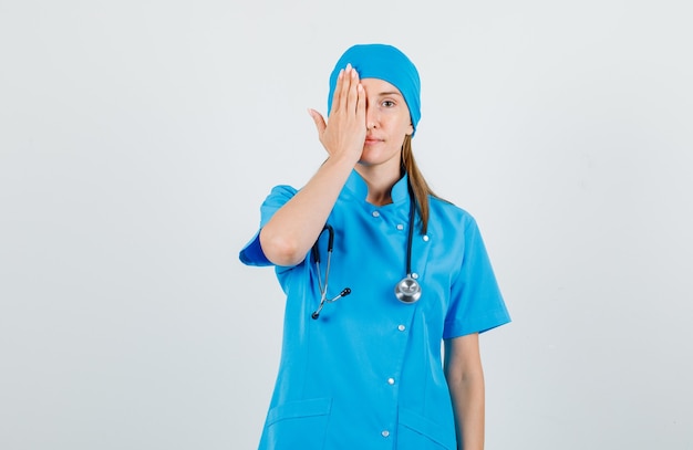 파란색 유니폼에 손으로 한쪽 눈을 덮고 긍정적 인 찾고 여성 의사. 전면보기.