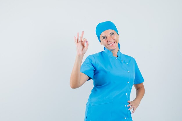 Female doctor doing ok gesture in blue uniform and looking confident , front view.