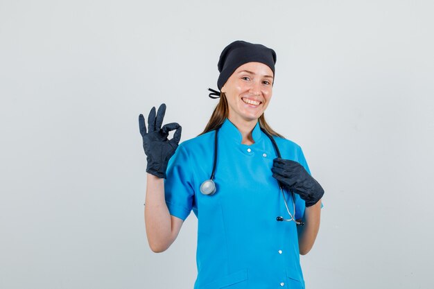 Female doctor doing ok sign with hand on stethoscope in uniform, gloves and looking glad