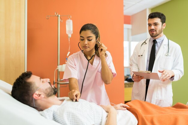 Female doctor examining sick patient with stethoscope while male doctor analyzing reports at hospital